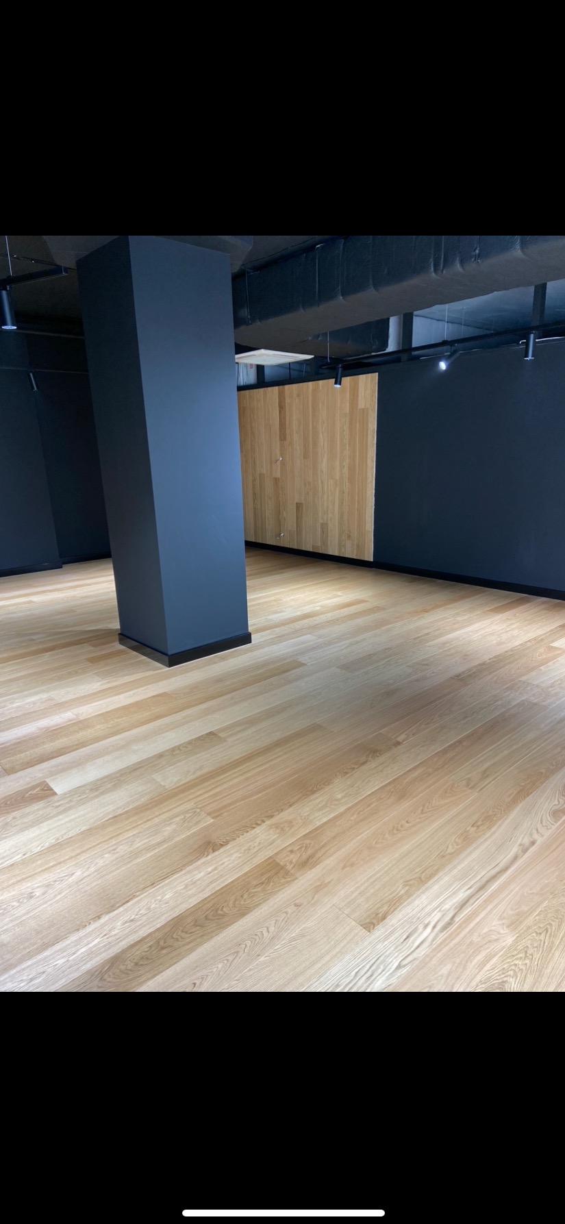 Jasna, drewniana podłoga z panelami ułożonymi ukośnie w przestronnym, nowoczesnym wnętrzu z ciemnymi ścianami i kolumną. Widoczne oświetlenie sufitowe.