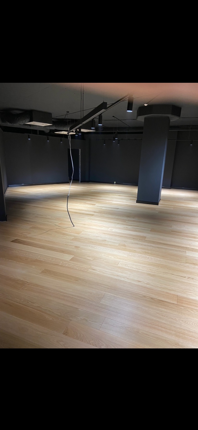 Jasne panele podłogowe w nowym pomieszczeniu z szarą ścianą i surowym oświetleniem, widoczne wiszące przewody.