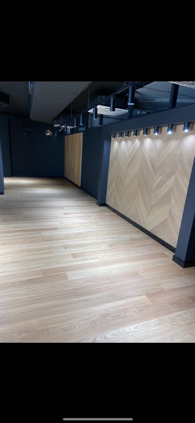 Jasna podłoga z paneli w Kamieńcu, ściana wykończona drewnianą mozaiką w jodełkę, sufit z czarnymi lampami punktowymi.