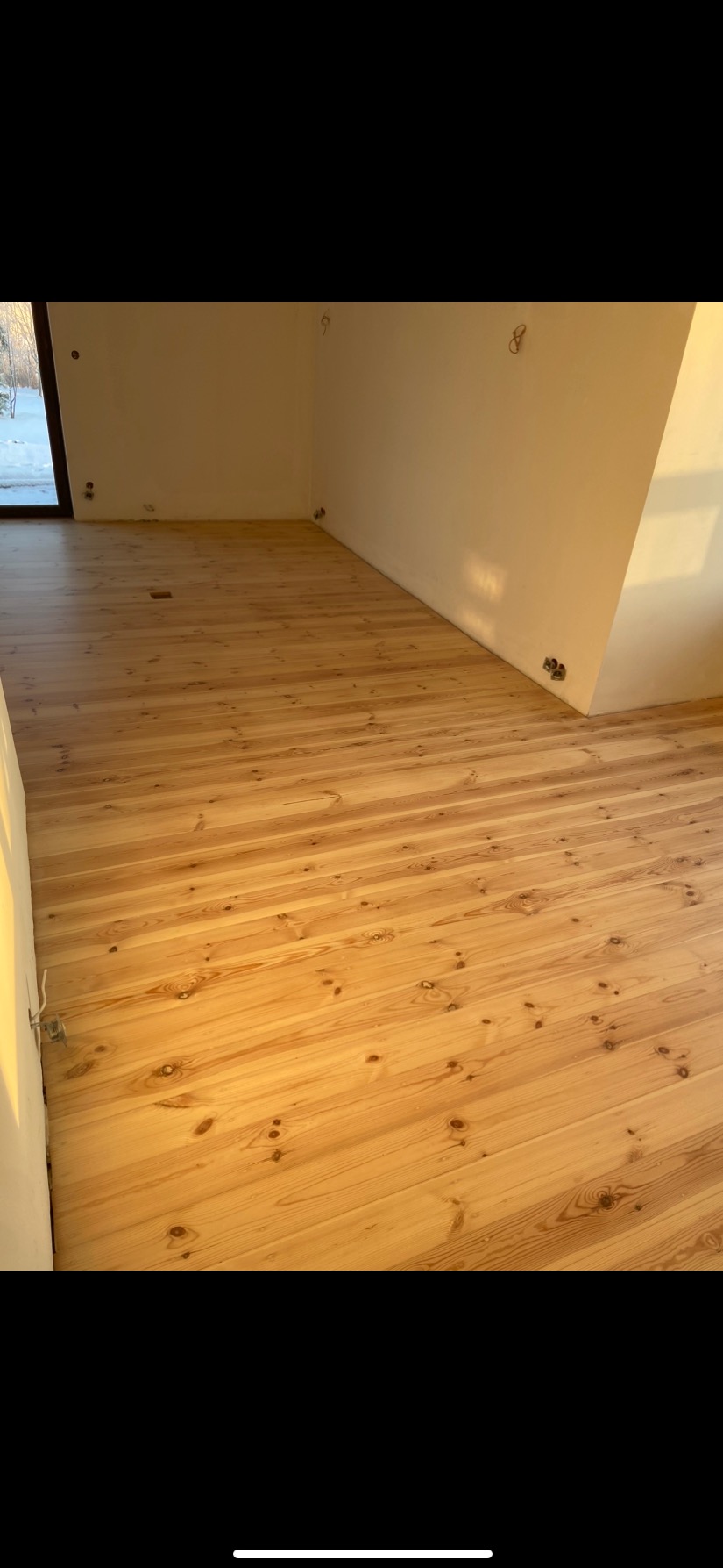 Jasna drewniana podłoga z widocznymi sękami, świeżo po renowacji, z widocznymi śladami po szlifowaniu w narożnikach pomieszczenia, przygotowana pod lakierowanie, z oknem w tle.