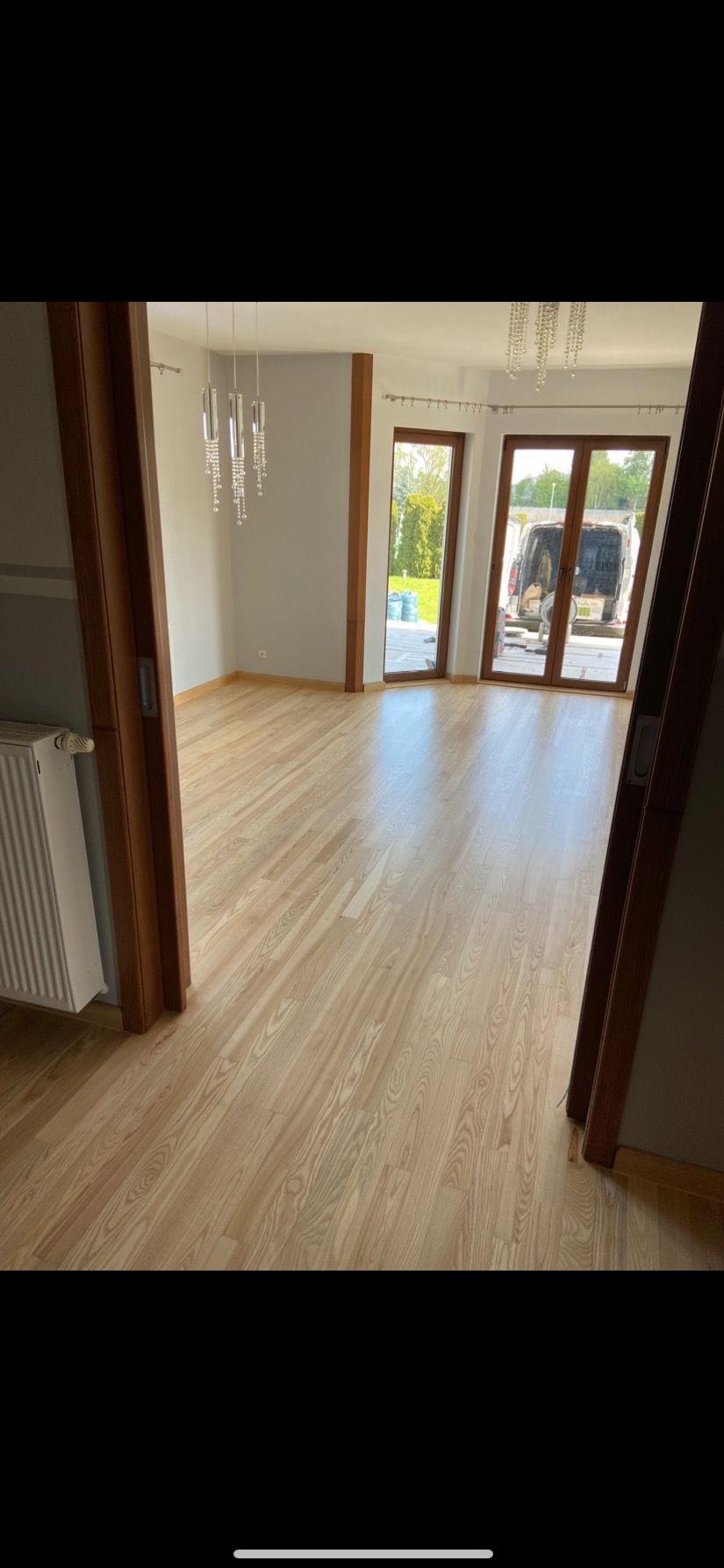 Jasna, świeżo ułożona podłoga z paneli drewnianych w pustym pokoju, widok przez otwarte drzwi na zewnątrz, gdzie stoi zaparkowany biały bus.