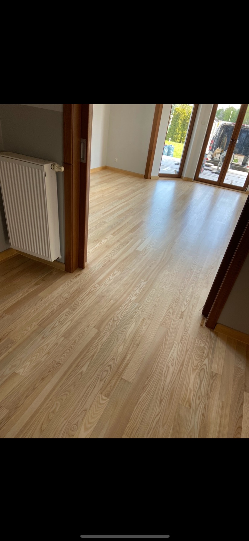 Jasny, drewniany parkiet ułożony w pustym pokoju, widoczny grzejnik i fragment okna z widokiem na zieleń.