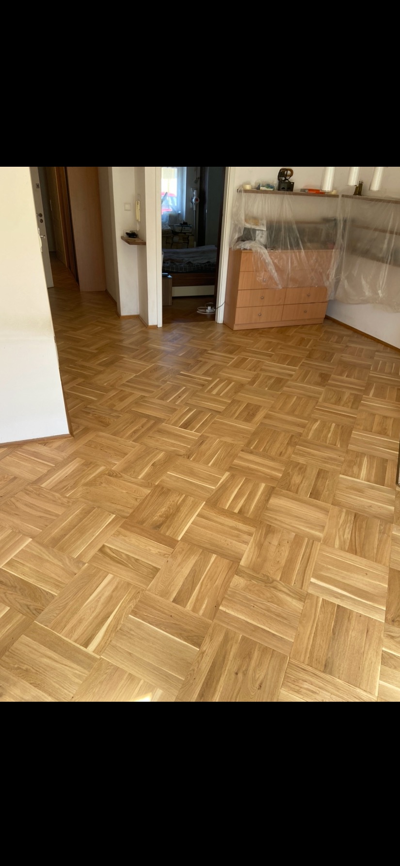 Parkiet ułożony w klasyczny wzór szachownicy, z widocznymi słojami drewna, w jasnym pomieszczeniu z meblami zabezpieczonymi folią ochronną.