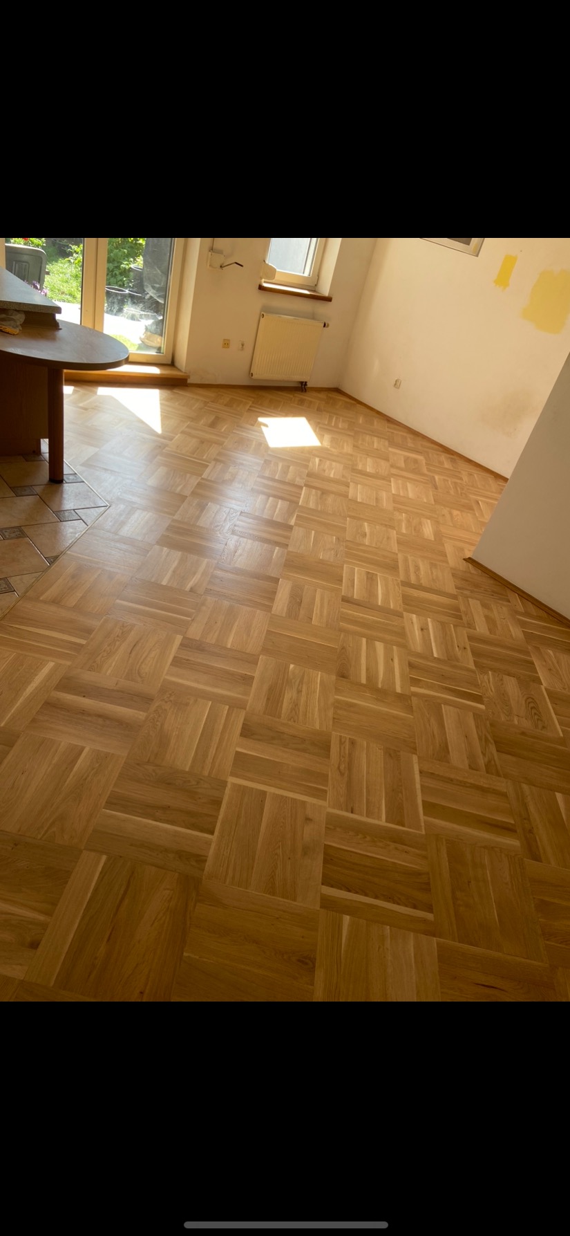 Parkiet ułożony w kwadraty, oświetlony popołudniowym słońcem wpadającym przez okno balkonowe, widoczne ślady po malowaniu na ścianie.