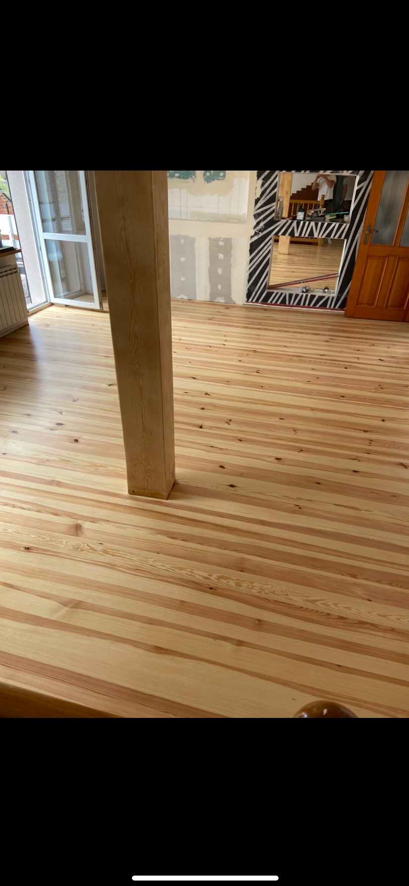 Jasne, świeżo położone deski podłogowe z widoczną drewnianą kolumną, w tle fragment ściany w trakcie remontu i lustro w czarno-białej ramie.