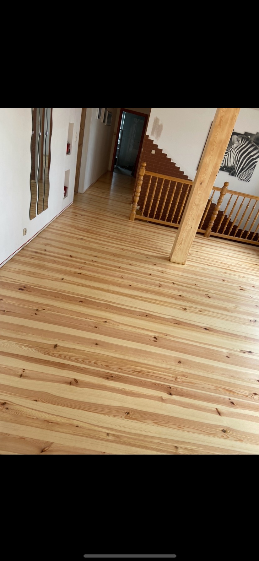 Jasna, drewniana podłoga z widocznymi słojami, świeżo po renowacji, z drewnianą balustradą schodów i lustrem na ścianie w tle.