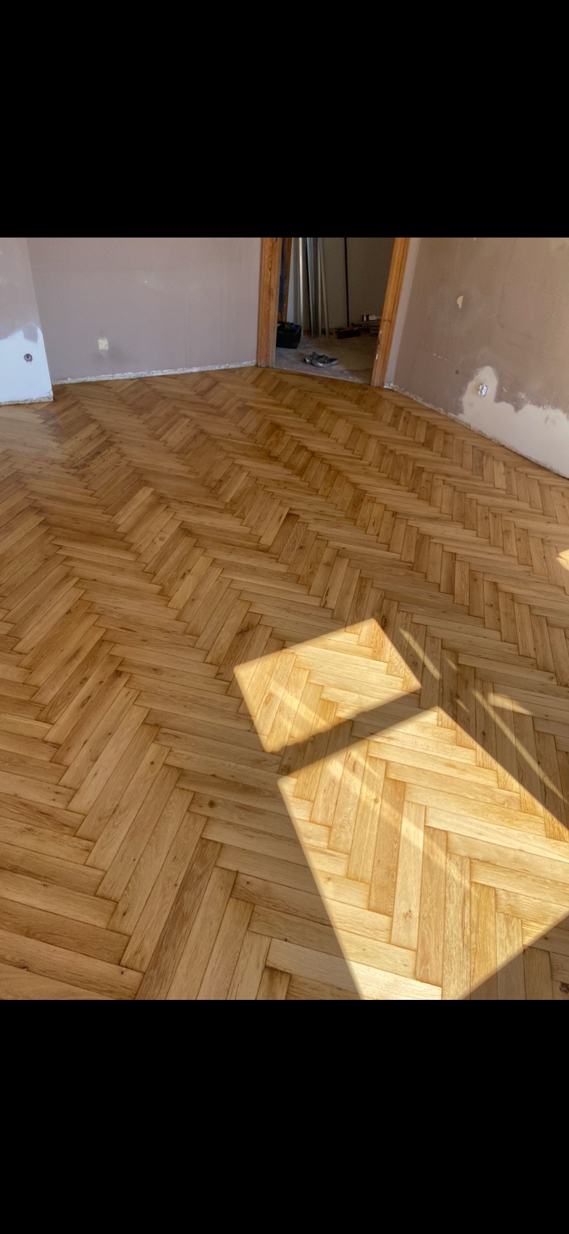 Ułożony parkiet w jodełkę w jasnym odcieniu drewna, z widocznymi promieniami słonecznymi padającymi na podłogę, ściany w trakcie remontu, widoczne ślady po łataniu ubytków.
