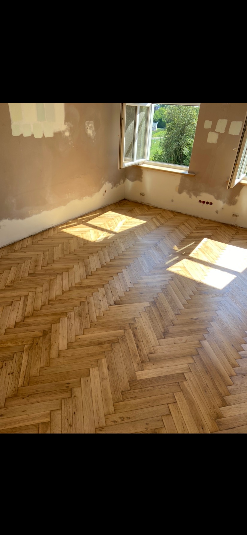 Nowo ułożona, jasna podłoga z parkietu w jodełkę w pokoju z przygotowanymi do malowania ścianami i otwartym oknem w słoneczny dzień.