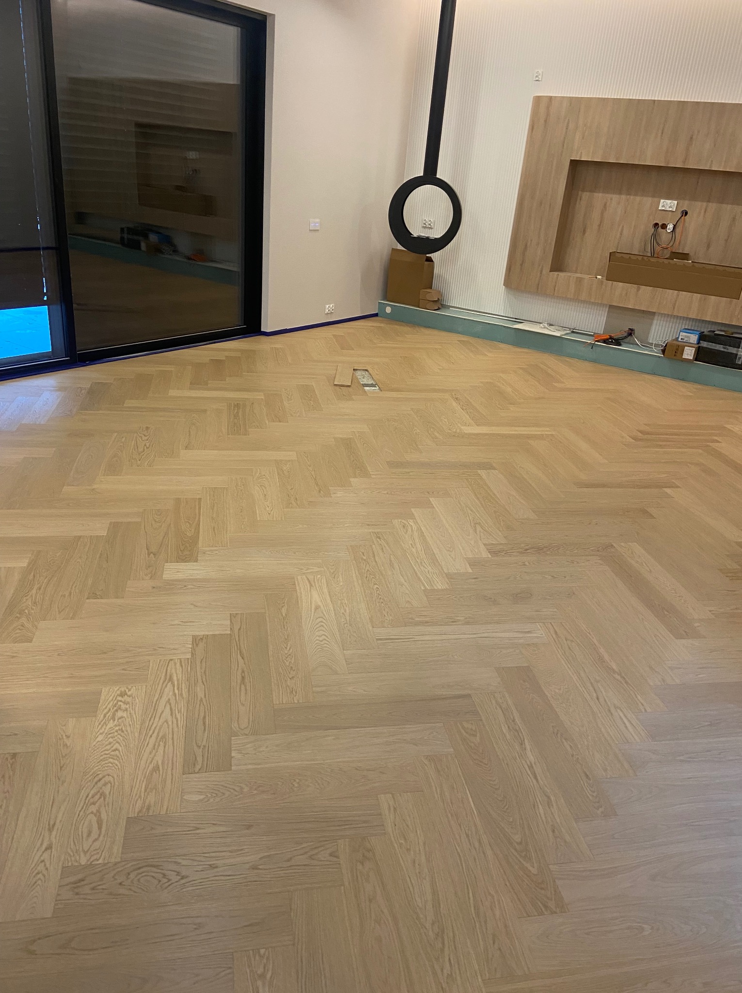 Jasna, nowa podłoga z parkietu ułożonego we wzór jodełki francuskiej, widoczne listwy przypodłogowe w trakcie montażu i zabezpieczenia taśmą malarską, w tle minimalistyczny kominek.