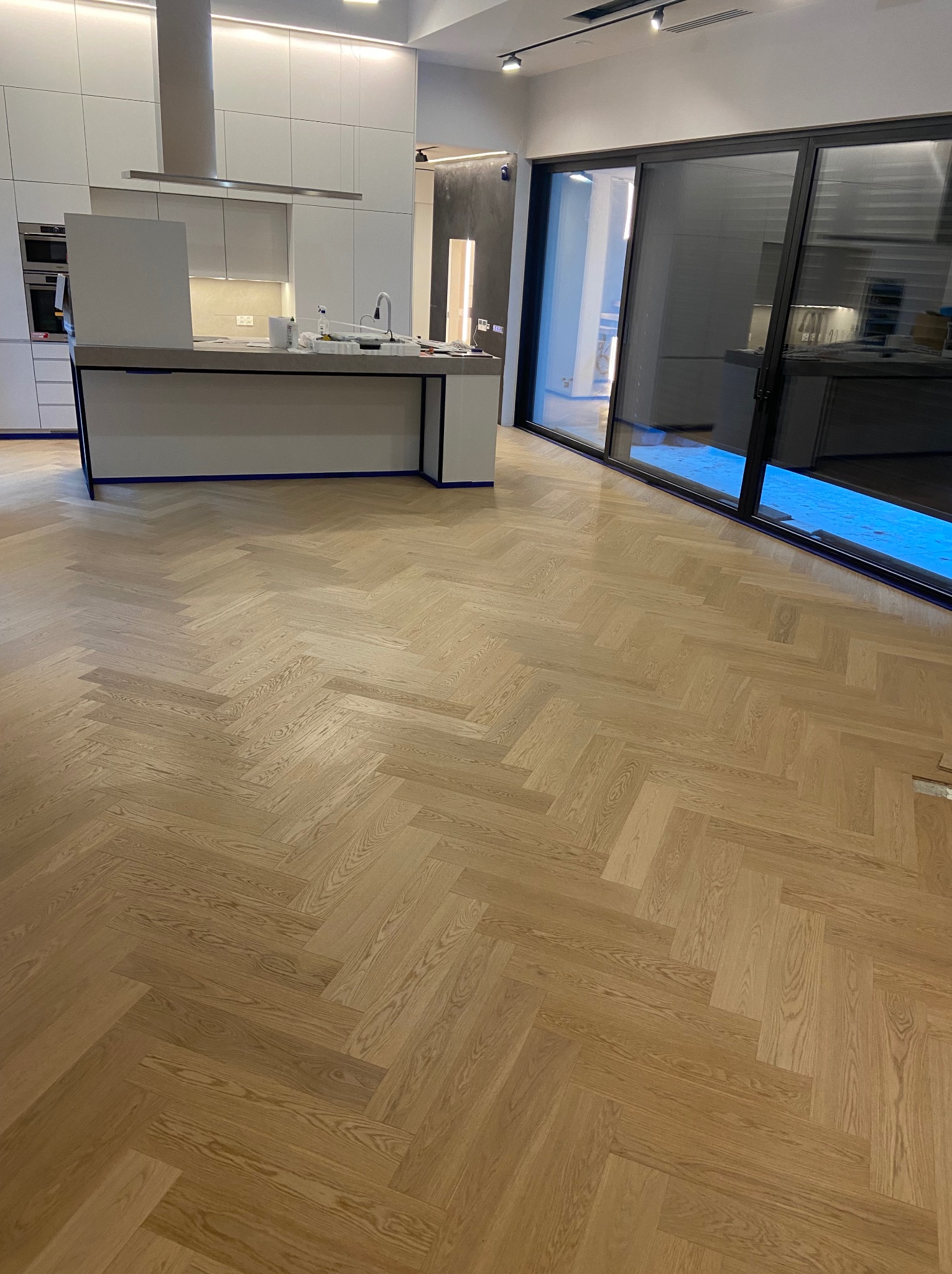 Jasne drewniane deski ułożone w jodełkę na podłodze w nowoczesnym wnętrzu z białą kuchnią i dużymi przesuwnymi drzwiami balkonowymi.