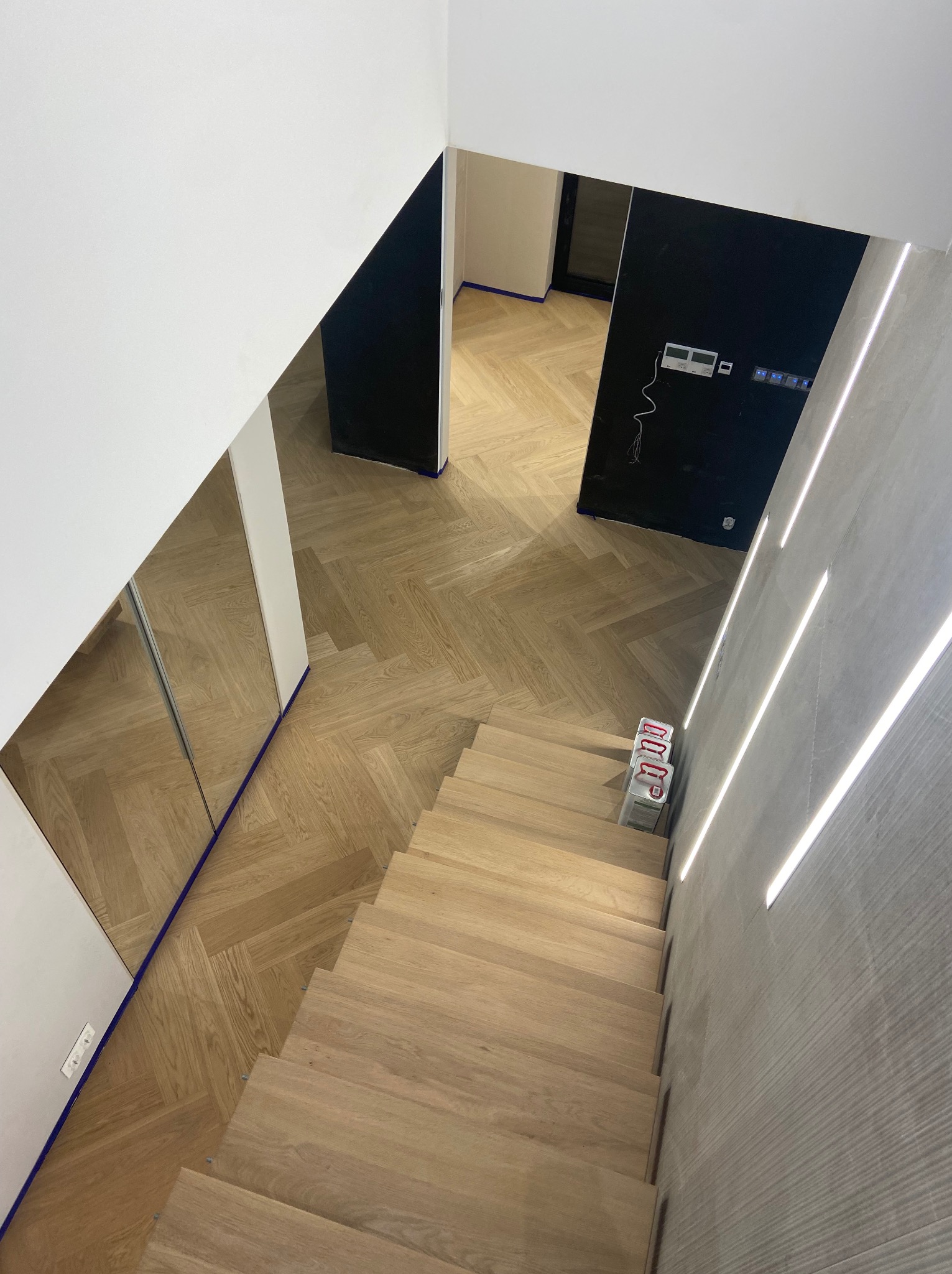 Widok z góry na drewniane schody i parkiet ułożony w jodełkę, zabezpieczone niebieską taśmą malarską, z widocznymi puszkami z lakierem na schodach i nowoczesnym oświetleniem w ścianie.