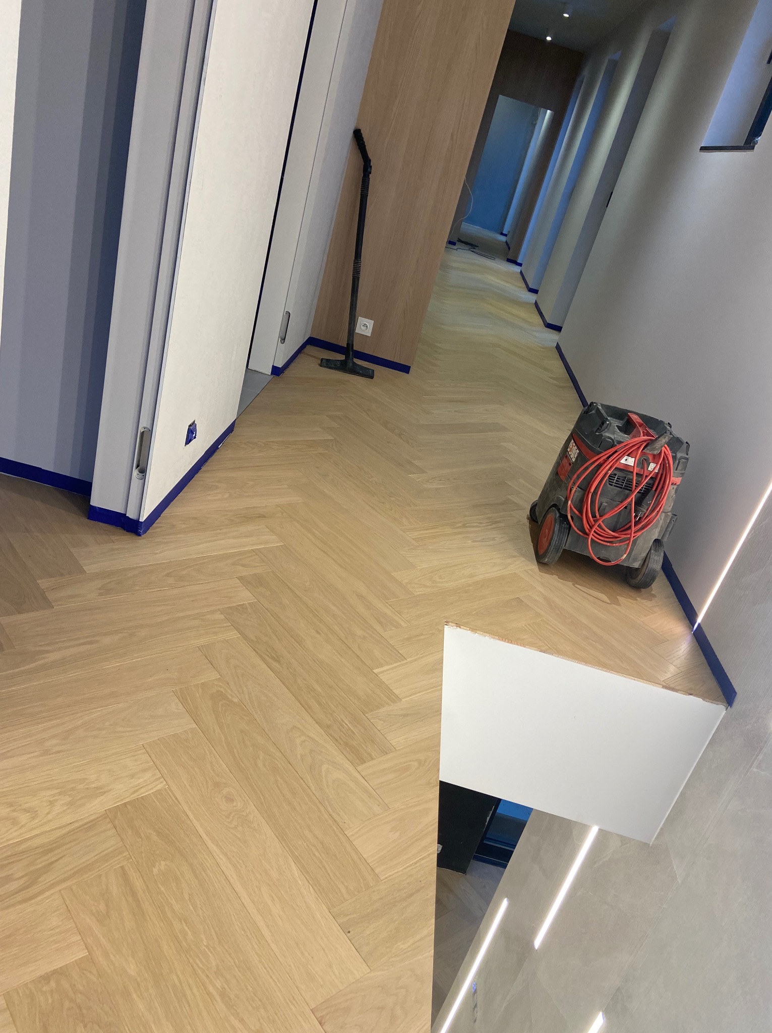 Nowo ułożona, jasna podłoga z parkietu w jodełkę, zabezpieczona niebieską taśmą malarską przy ścianach i drzwiach; odkurzacz przemysłowy z czerwonym kablem stoi w rogu korytarza.