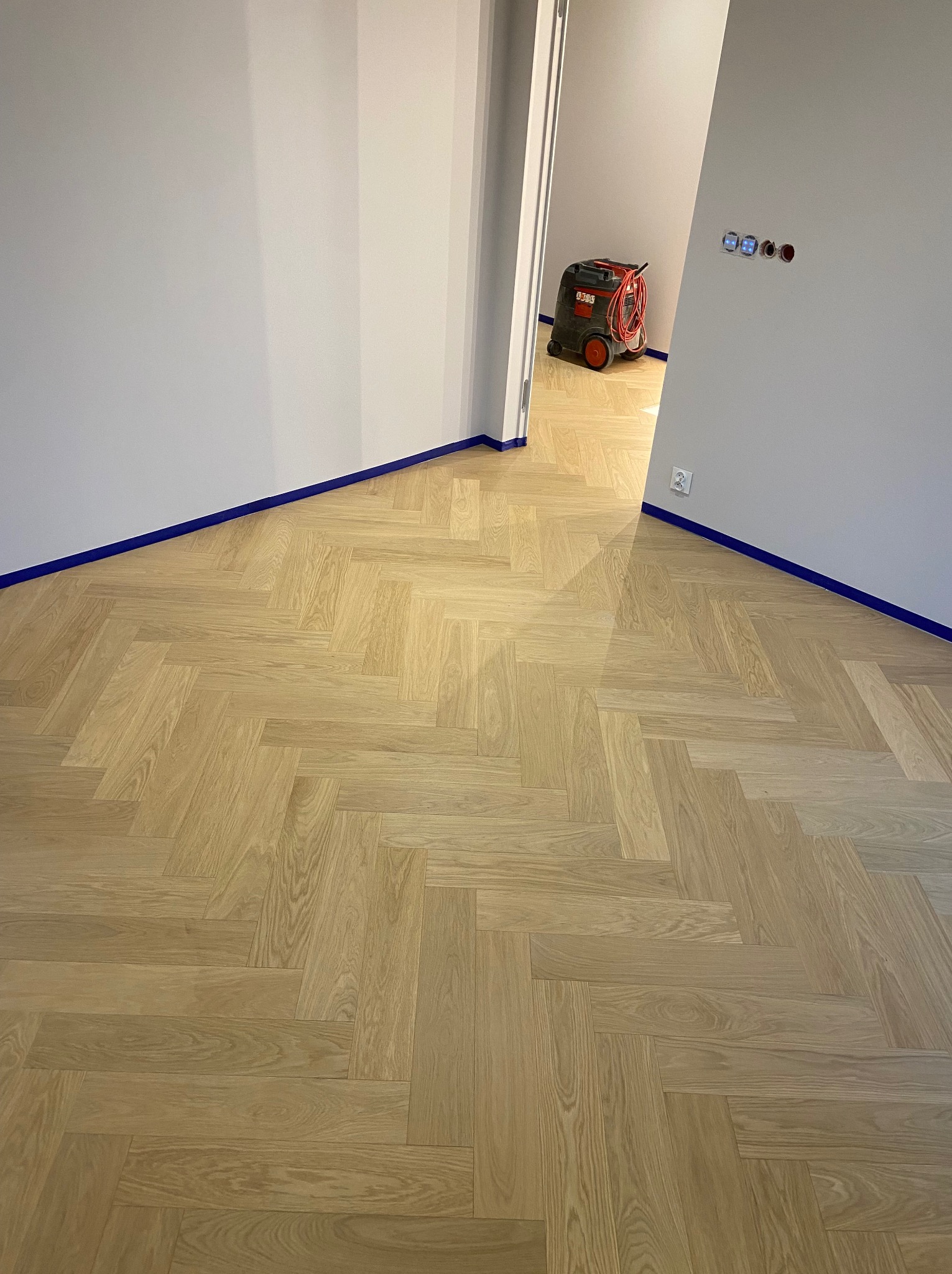 Jasny parkiet ułożony w jodełkę francuską, ściany zabezpieczone niebieską taśmą malarską, w tle widoczny odkurzacz przemysłowy