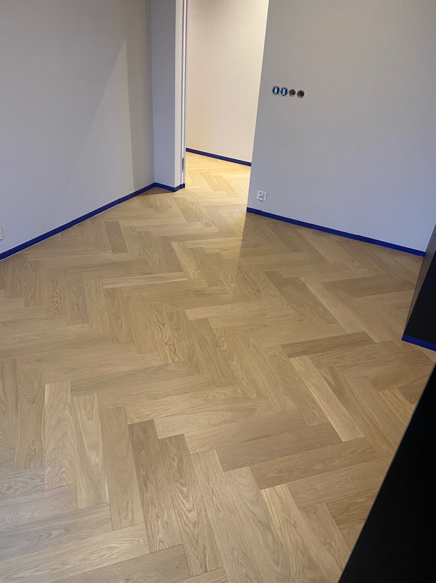 Ułożony parkiet w jodełkę, zabezpieczony niebieską taśmą malarską przy ścianach w szarym odcieniu, widoczne gniazdko elektryczne i puszki instalacyjne bez osprzętu.