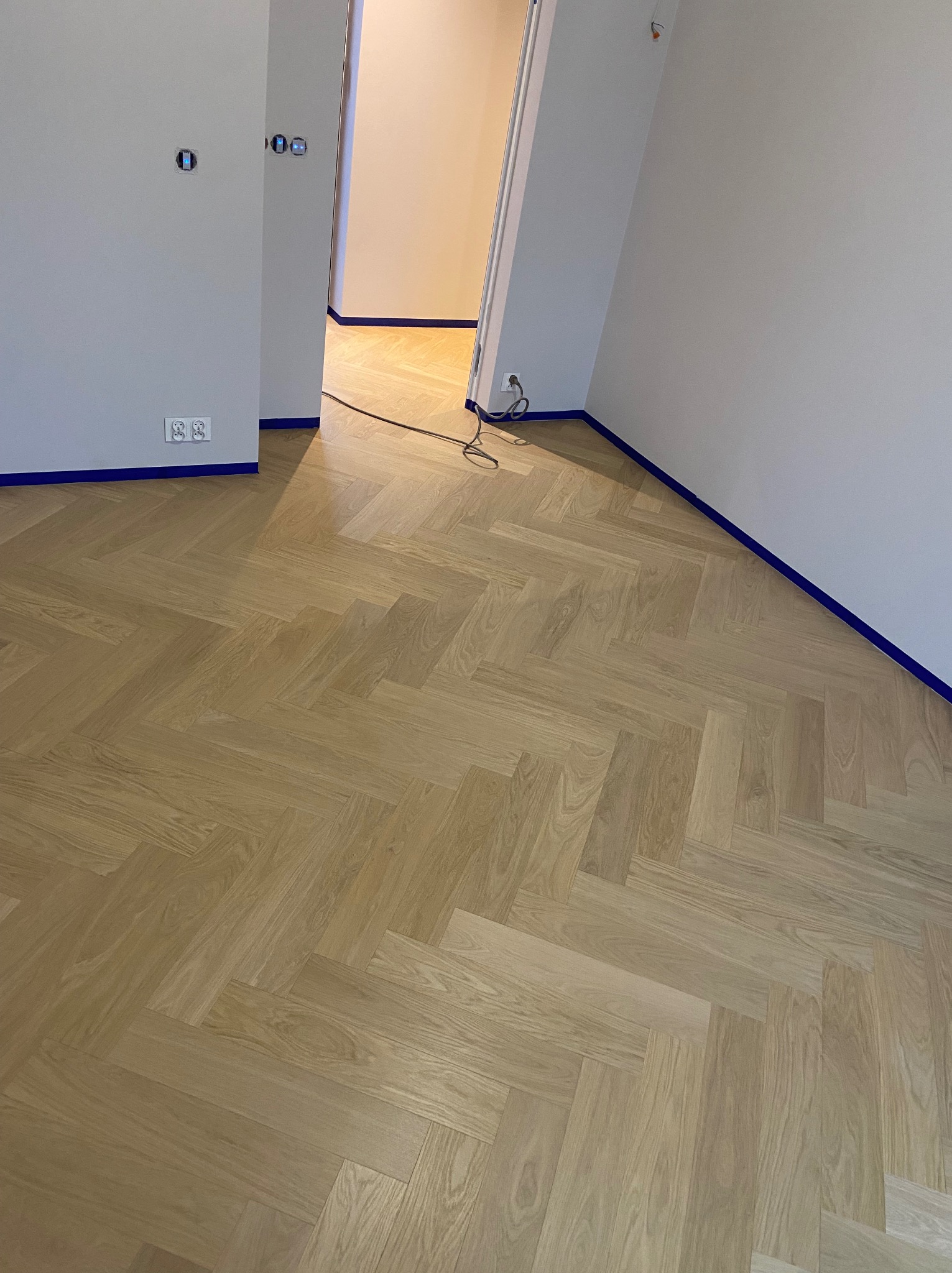 Nowo ułożona, jasna podłoga parkietowa w jodełkę francuską, zabezpieczona niebieską taśmą malarską przy ścianach w trakcie remontu.