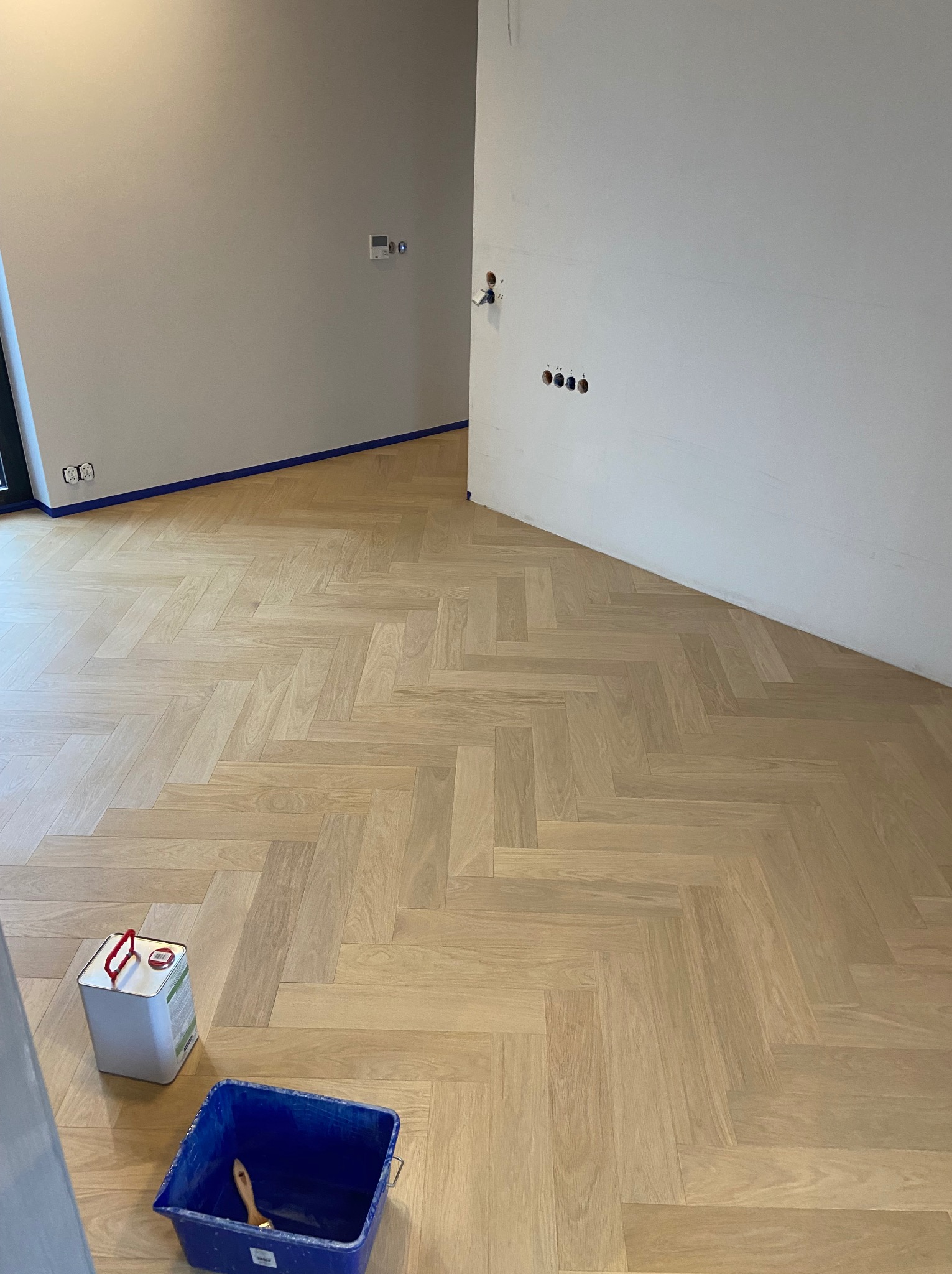 Jasna, nowa podłoga z parkietu ułożonego w jodełkę francuską, zabezpieczona niebieską taśmą malarską przy ścianach; obok puszka lakieru i kuweta z pędzlem.