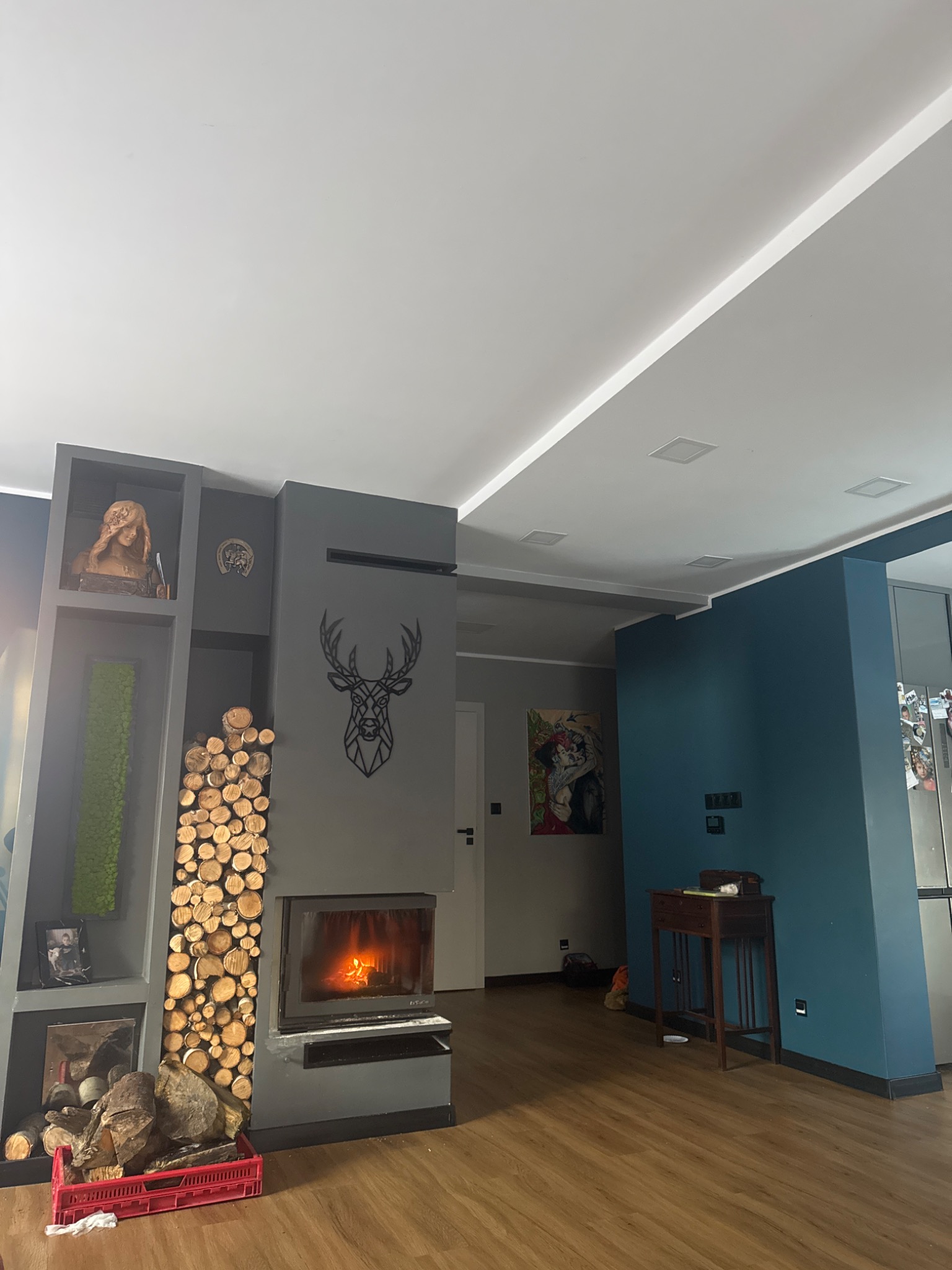 Nowoczesny salon z szarą ścianą, kominkiem z dekoracją jelenia, drewnem ułożonym obok, drewnianą podłogą i sufitem z wbudowanym oświetleniem