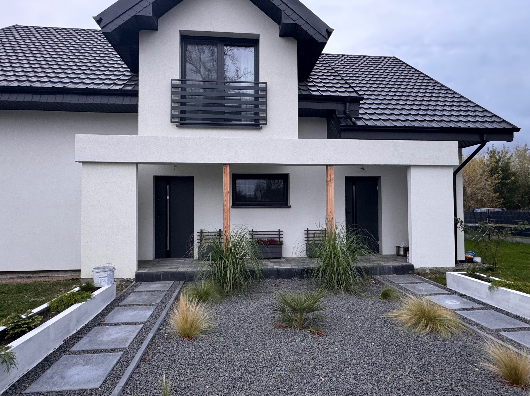 Nowoczesny dom jednorodzinny z białymi ścianami, czarnym dachem i stolarką okienną, otoczony szarym żwirem i ozdobnymi trawami, widok na elewację frontową z dwoma wejściami.
