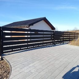 STALVA Piotr Sadkowski - Nowoczesna, pozioma, czarna brama wjazdowa z przesuwnym mechanizmem, widoczna na tle domu jednorodzinnego i brukowanego podjazdu w słoneczny dzień.