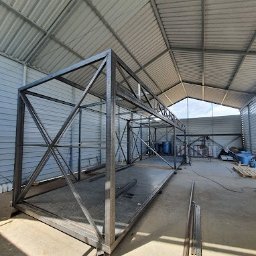 STALVA Piotr Sadkowski - Szkielet metalowej konstrukcji w hali, widoczne spawane profile tworzące ramę i kratownice, surowy stan przed wykończeniem, stalowa podstawa z kratą.