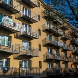 Fasada budynku z licznymi balkonami z metalowymi balustradami, niektóre z antenami satelitarnymi, w słoneczny dzień.