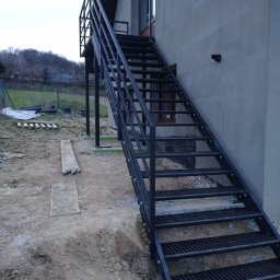 STALVA Piotr Sadkowski - Zewnętrzne schody metalowe przy budynku, z ażurowymi stopniami i barierką. Widoczne mocowania do ściany. Tło to ziemia i zieleń, pochmurne niebo.