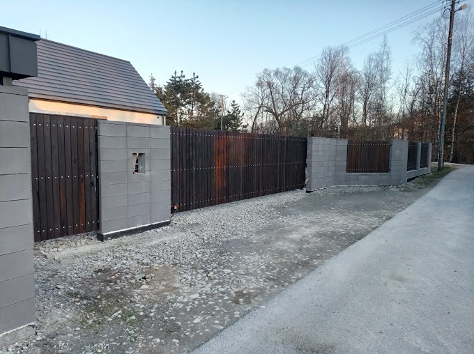 Nowoczesna posesja z drewnianą bramą wjazdową i ogrodzeniem, osadzone w betonowych słupkach, widok z zewnątrz na żwirowy podjazd.