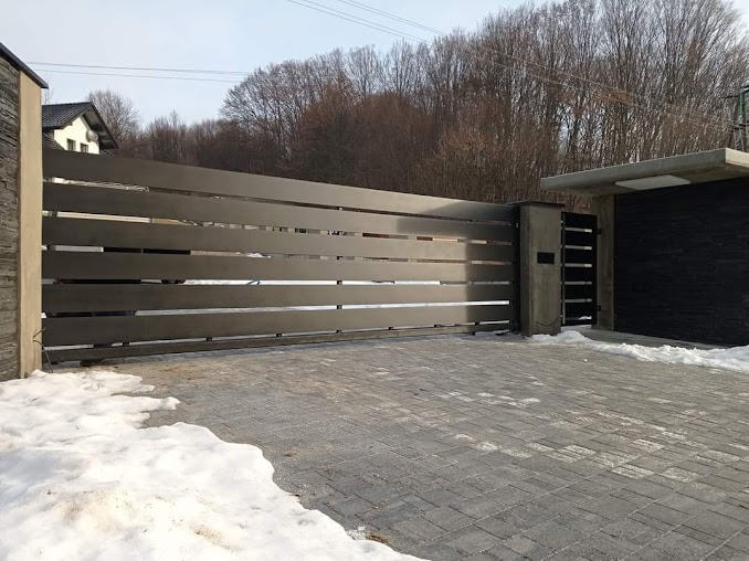 Nowoczesna brama wjazdowa z poziomych, metalowych profili w kolorze szarości, zamontowana na posesji z kostką brukową i resztkami śniegu.
