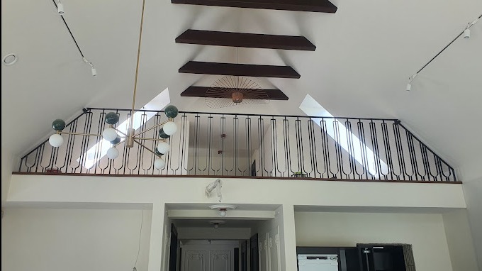 Czarna, metalowa barierka na antresoli w jasnym wnętrzu z drewnianymi belkami stropowymi i nowoczesnym żyrandolem. W tle widoczne okno dachowe wpuszczające światło.