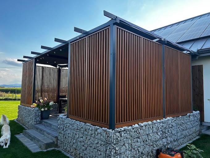 Nowoczesna pergola z drewnianymi lamelami i gabionową podstawą, z widocznym robotem koszącym i psem na trawniku.