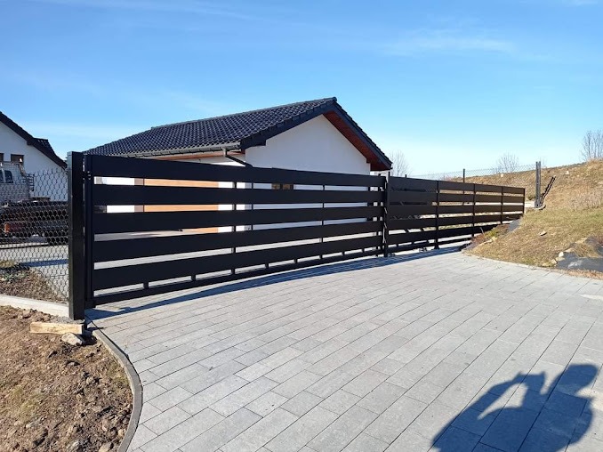 Nowoczesna, pozioma, czarna brama wjazdowa z przesuwnym mechanizmem, widoczna na tle domu jednorodzinnego i brukowanego podjazdu w słoneczny dzień.