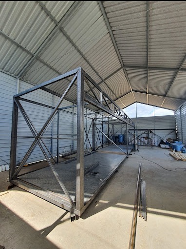 Szkielet metalowej konstrukcji w hali, widoczne spawane profile tworzące ramę i kratownice, surowy stan przed wykończeniem, stalowa podstawa z kratą.