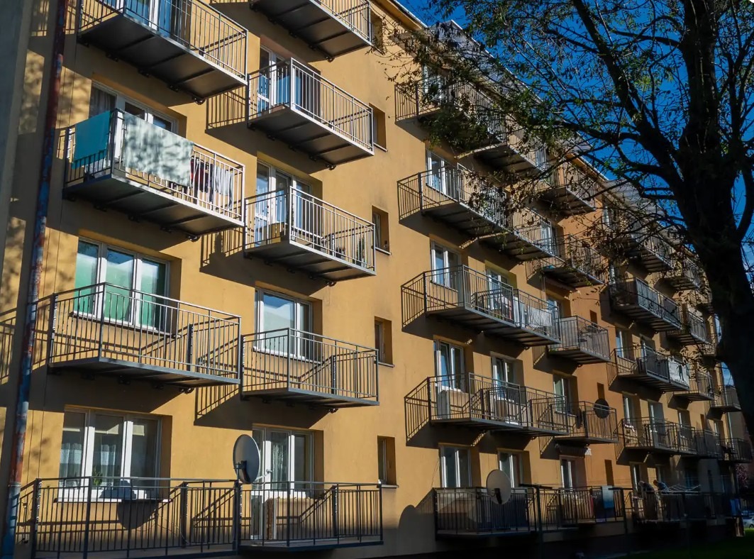 Fasada budynku z licznymi balkonami z metalowymi balustradami, niektóre z antenami satelitarnymi, w słoneczny dzień.