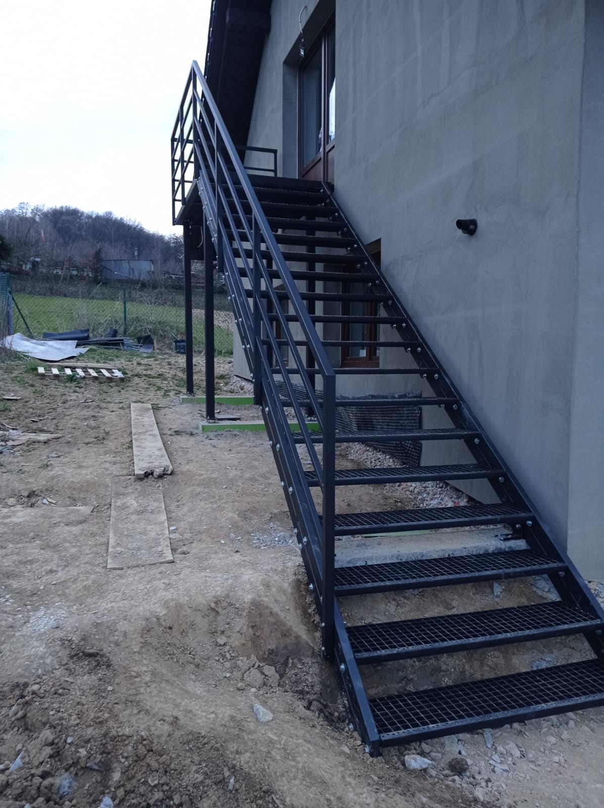 Zewnętrzne schody metalowe przy budynku, z ażurowymi stopniami i barierką. Widoczne mocowania do ściany. Tło to ziemia i zieleń, pochmurne niebo.