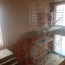 Praca na budowie: murarz na rusztowaniu tynkuje ścianę z cegły, widoczna część ściany już pokryta tynkiem, surowy betonowy strop