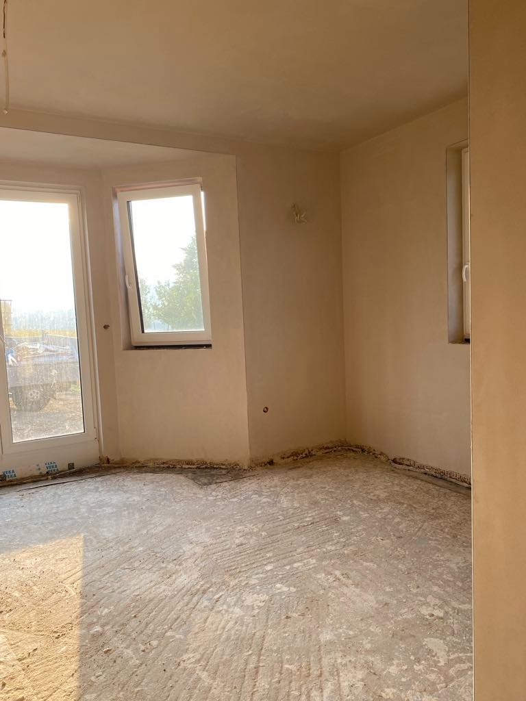 Surowe wnętrze po nałożeniu tynku maszynowego: widoczne ślady po tynku na podłodze, okna z widokiem na zewnątrz, ściany pokryte świeżym tynkiem w odcieniach beżu.
