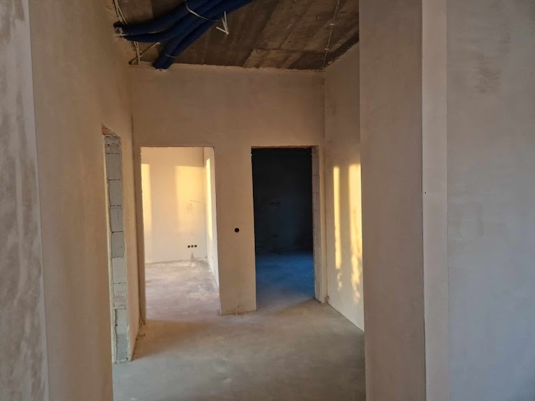 Wnętrze w trakcie tynkowania maszynowego: ściany pokryte świeżym tynkiem, widoczne instalacje pod sufitem i surowa rama drzwiowa.