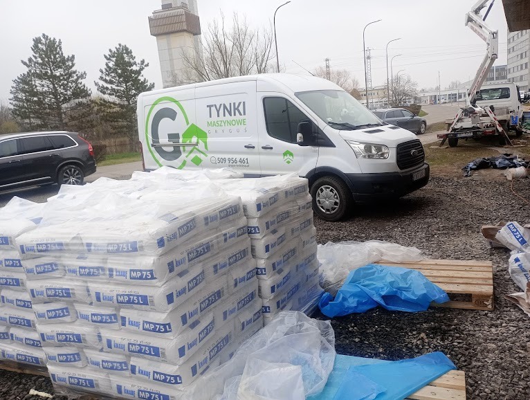 Stosy worków zaprawy gipsowej MP 75 L na palecie, z firmowym busem w tle, widoczny podnośnik koszowy oraz niebieskie folie ochronne na żwirowym podłożu.