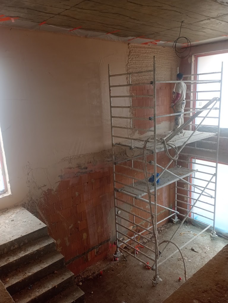 Praca na budowie: murarz na rusztowaniu tynkuje ścianę z cegły, widoczna część ściany już pokryta tynkiem, surowy betonowy strop