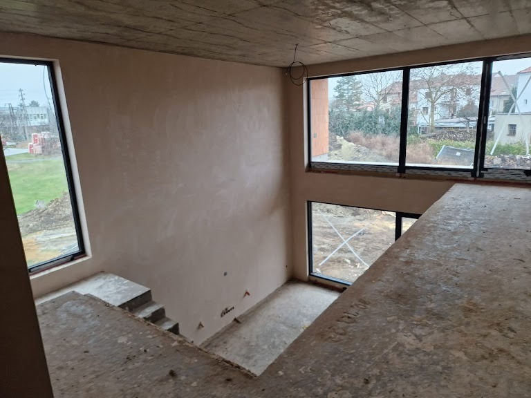 Wnętrze surowego domu po nałożeniu gładzi gipsowej na ściany, widoczne okna z czarnymi ramami i surowy betonowy sufit z wystającymi kablami.