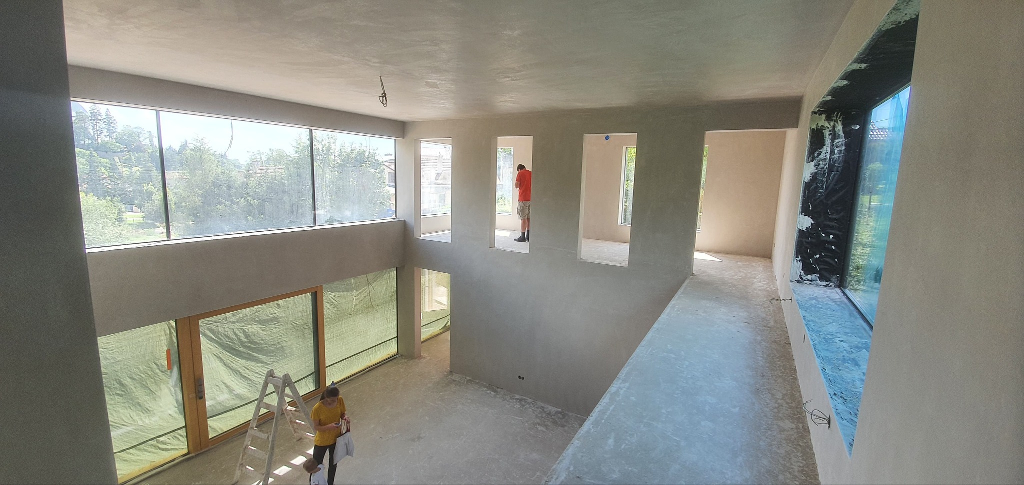 Wnętrze domu z wysokimi oknami po nałożeniu gładzi, widoczna drabina, kobieta i mężczyzna w oddali.