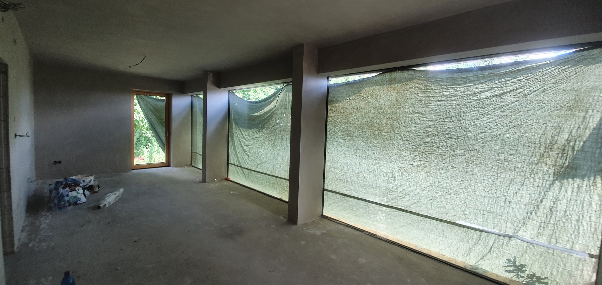 Wnętrze surowego pomieszczenia po nałożeniu tynków, z oknami zasłoniętymi zieloną plandeką, widoczne ślady po materiałach budowlanych na podłodze i ścianach.