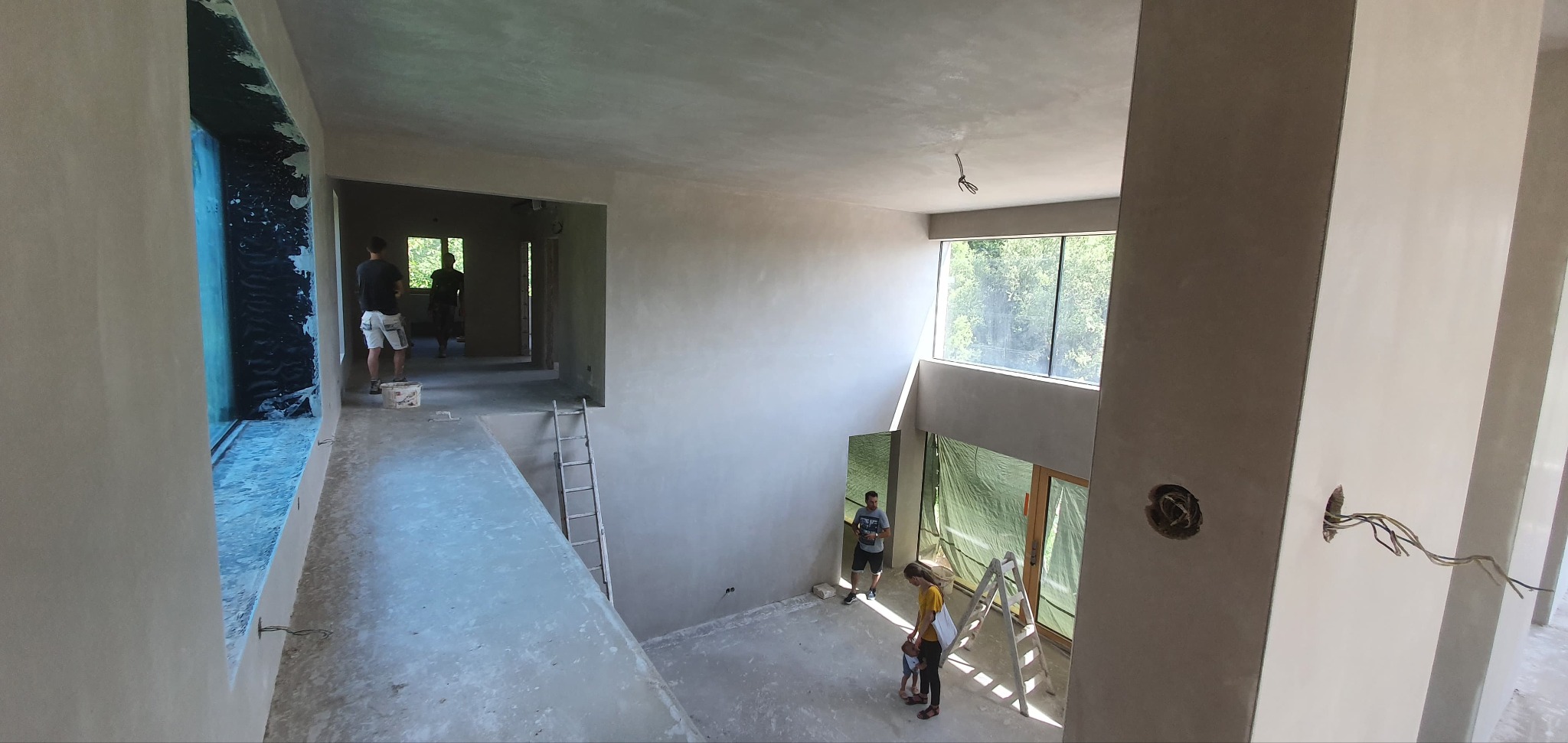 Wnętrze dwupoziomowego domu w trakcie prac wykończeniowych, widoczne świeżo położone tynki na ścianach i suficie, drabina oparta o ścianę i okno zabezpieczone folią, osoby w oddali.