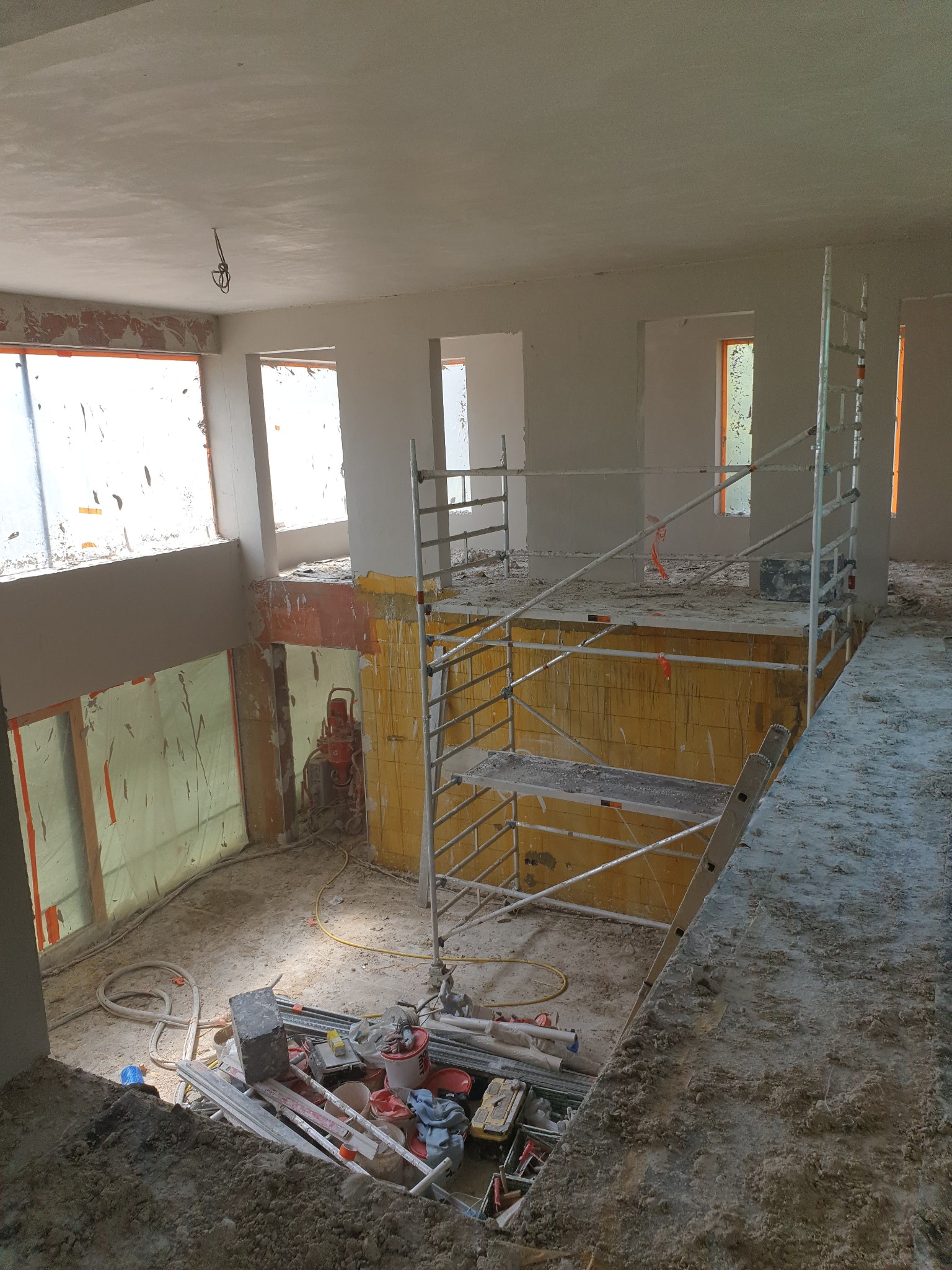 Wnętrze budynku w trakcie tynkowania maszynowego, widoczne rusztowanie, agregat tynkarski, zabezpieczone okna folią, świeżo nałożony tynk na suficie i ścianach, oraz bałagan budowlany na podłodze.