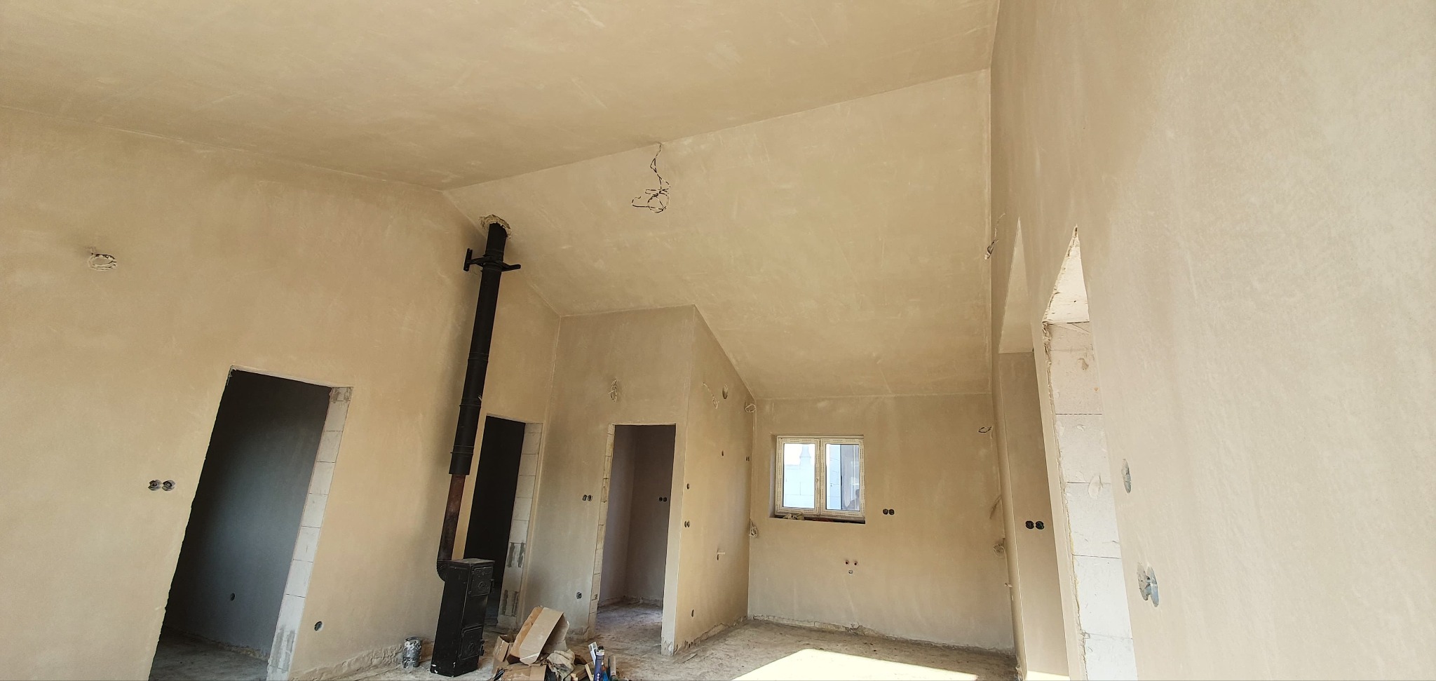 Wnętrze domu po nałożeniu jasnych tynków, widoczny piec typu koza z czarną rurą, niezabudowane otwory drzwiowe i okno z widokiem na zewnątrz, ślady po instalacji elektrycznej.
