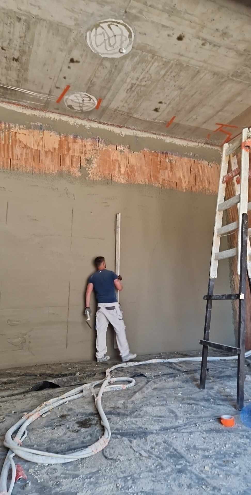 Aplikacja gładzi na ścianie z cegły, widoczne przewody i drabina, surowy stan budynku.