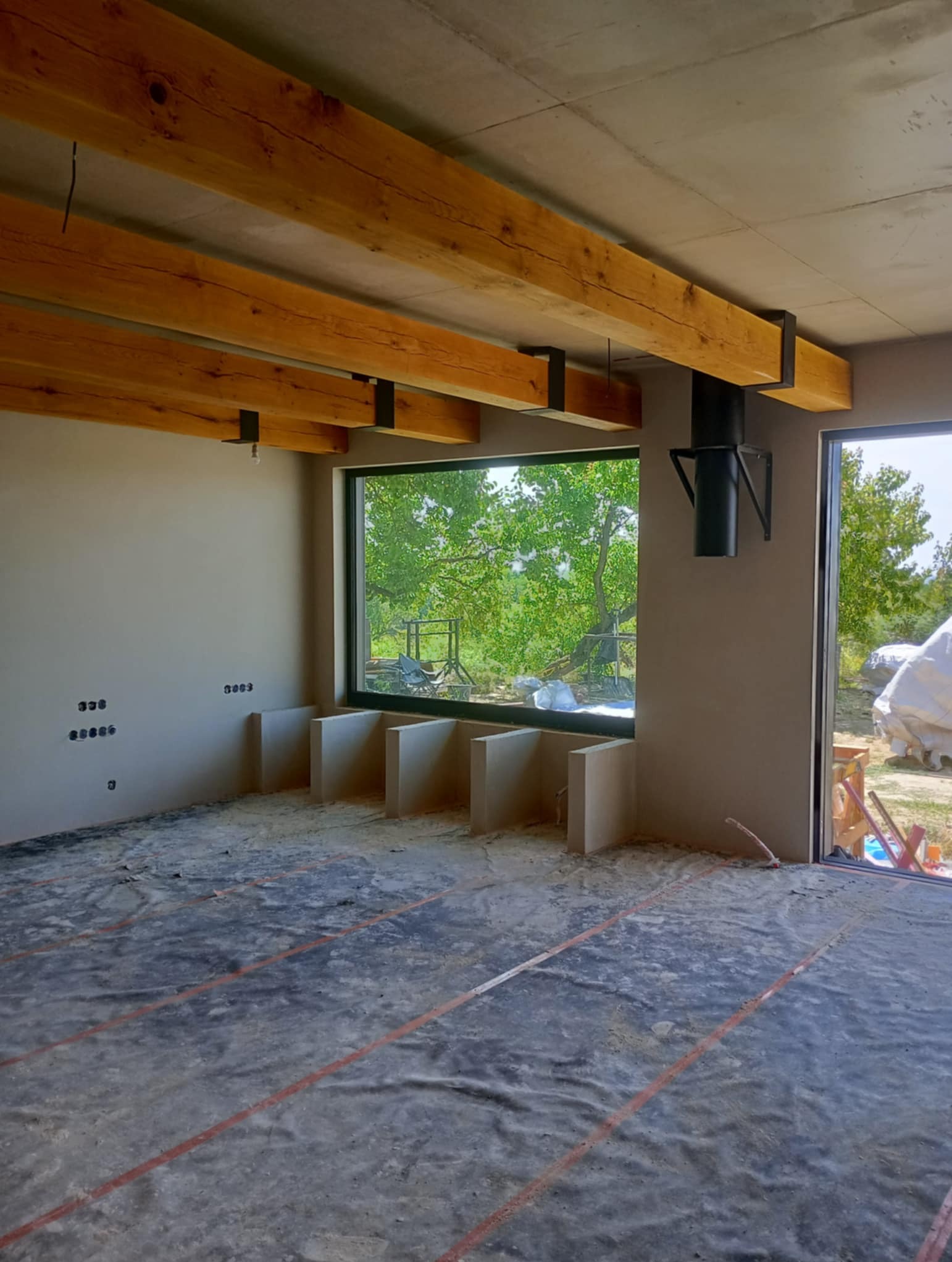 Surowe wnętrze z drewnianymi belkami stropowymi i przygotowaną podłogą pod ogrzewanie podłogowe, widok na zieleń za oknem.