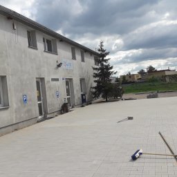S&L masonry Sylwia Dreas - Nowo ułożona, jasnoszara kostka brukowa przed budynkiem biurowym z oznaczeniami parkingowymi, widoczna szczotka i listwa do rozprowadzania piasku, pochmurne niebo.