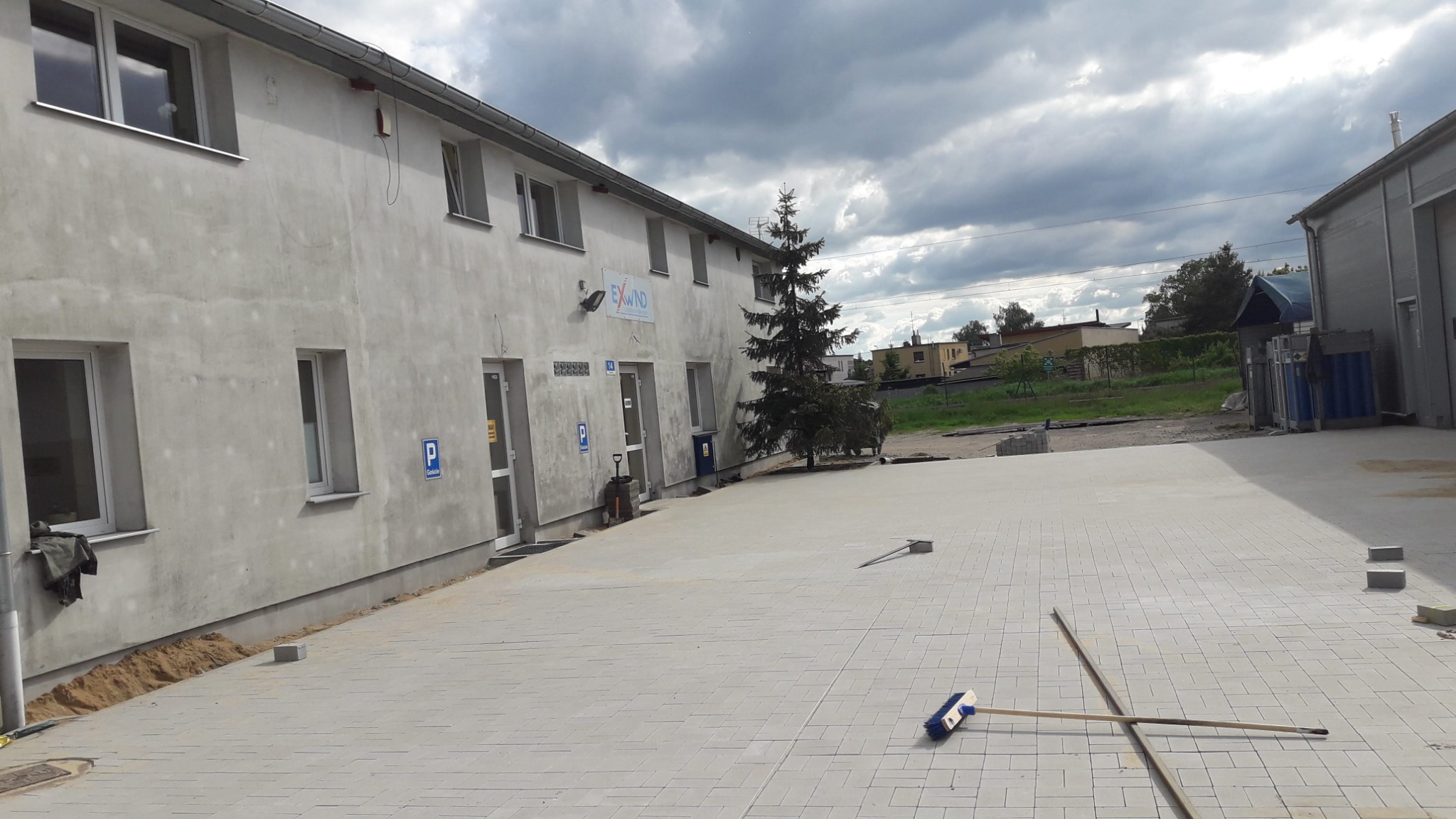 Nowo ułożona, jasnoszara kostka brukowa przed budynkiem biurowym z oznaczeniami parkingowymi, widoczna szczotka i listwa do rozprowadzania piasku, pochmurne niebo.