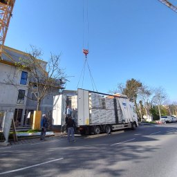 ECSI SPEDYCJA SPÓŁKA Z OGRANICZONĄ ODPOWIEDZIALNOŚCIĄ - Rozładunek materiałów budowlanych z naczepy ciężarówki za pomocą dźwigu na tle budynku w trakcie budowy, widoczni pracownicy w kaskach.